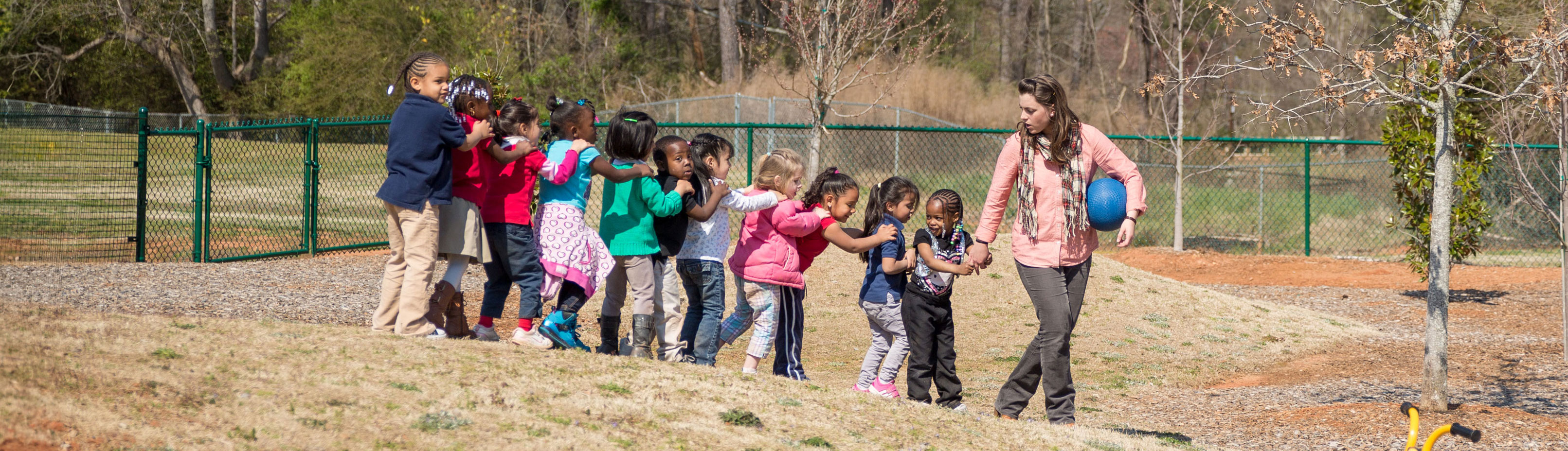 A teacher leads a large group of children in a single file line outdoors.  The children have their hands on each other's shoulders