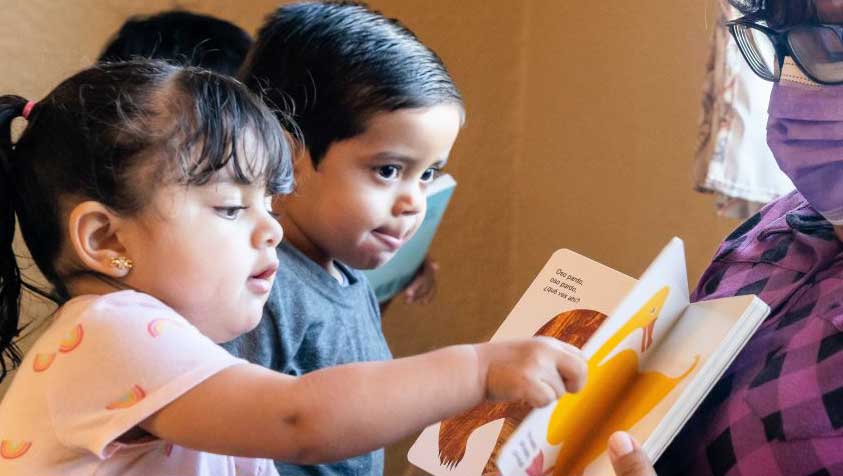 Two toddlers look at a picture book while a caregiver holds it up.  The toddler in the foreground is flipping the page over.