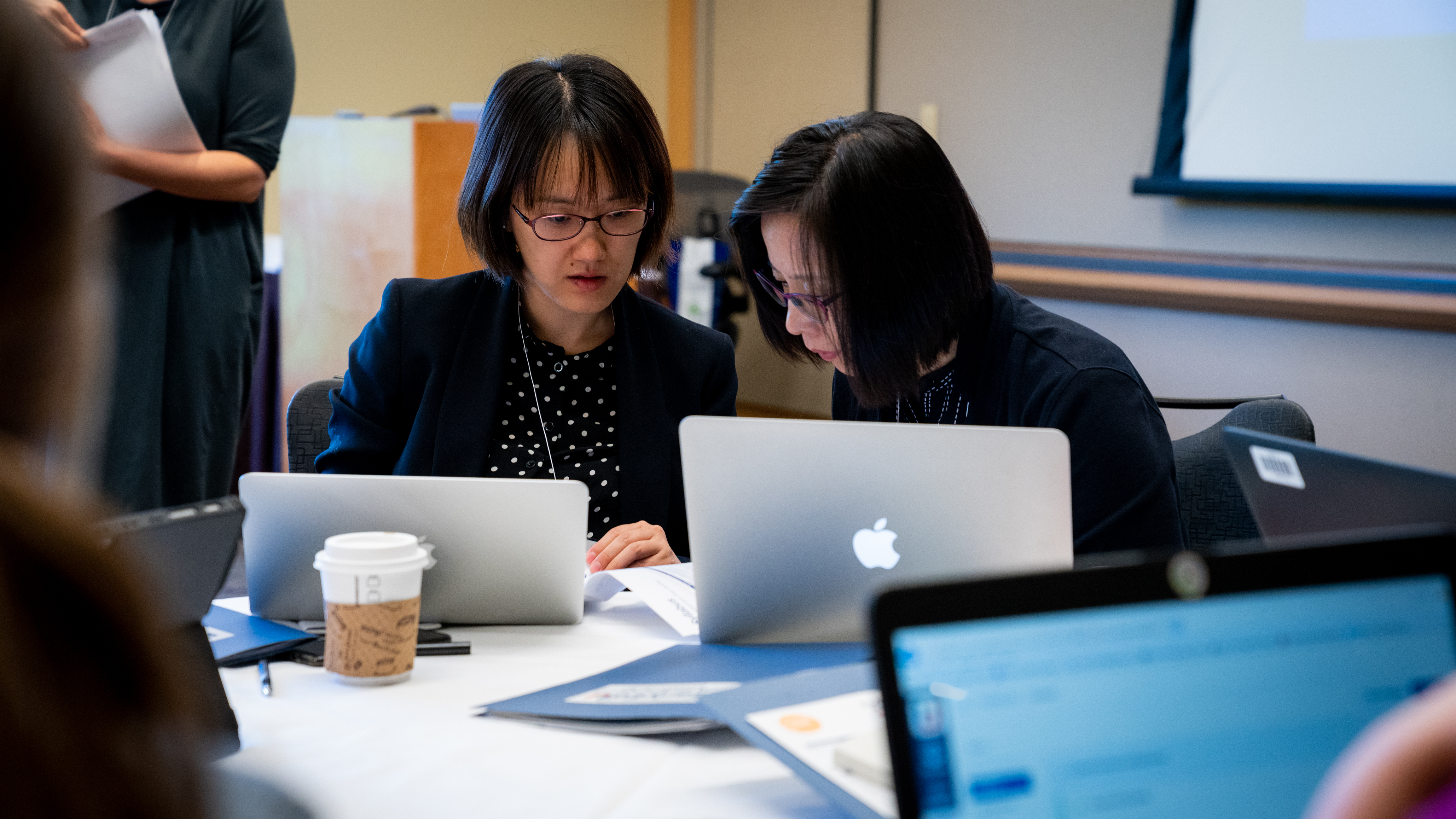 Two adults sitting at a table with laptops opened are reviewing a piece of paper. They are in a conference setting.